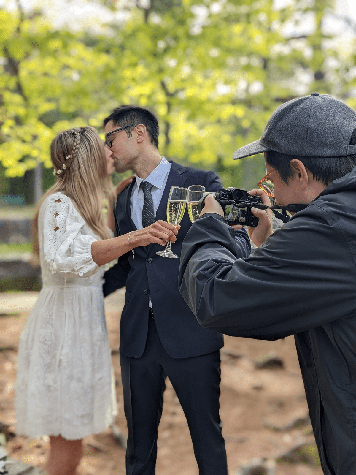 A photographer (Tyler Colburne) takes a picture of a person in a white dress and a person in a suit kissing while cheersing glasses.