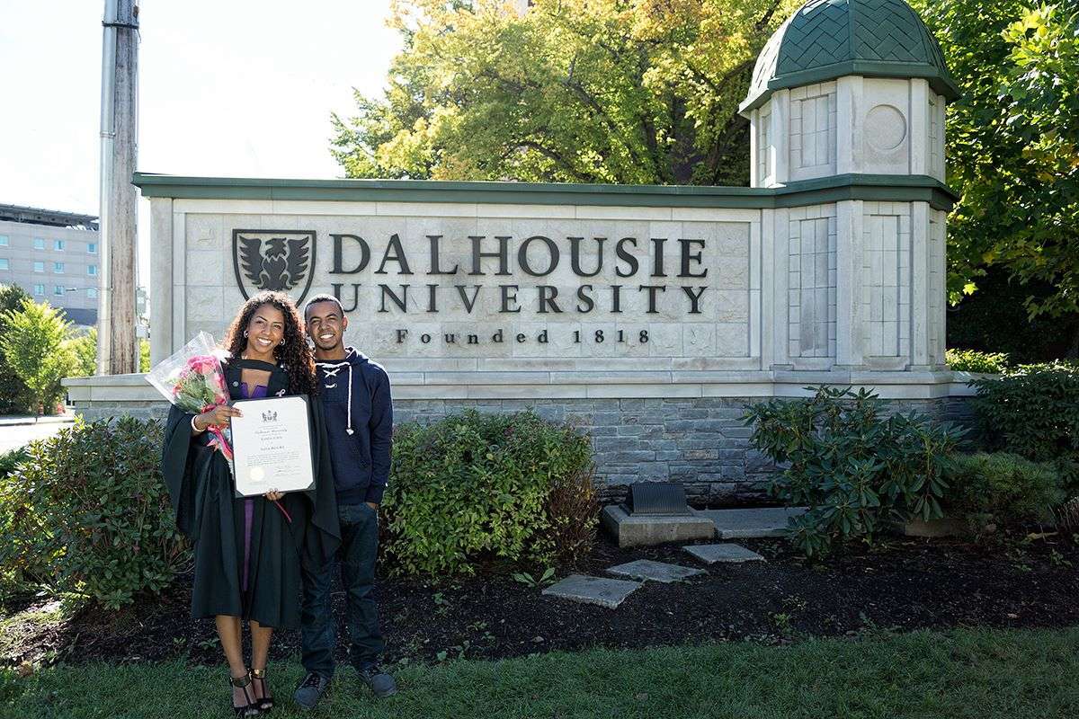 A photo of Ashley Hill in her graduation gown standing next to a person in front of the Dalhousie University sign