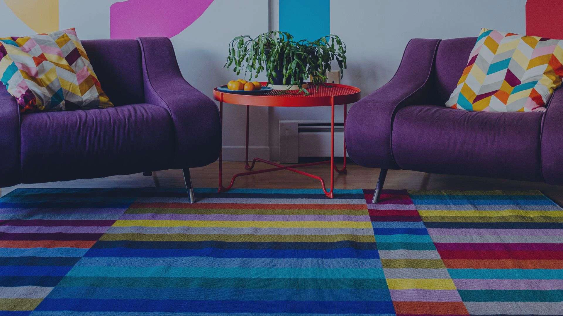 Photo of a colorful carpet with two purple chairs and a table with a plant on it.