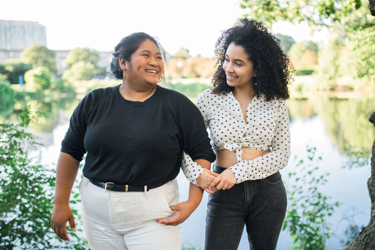 A photo of two people linked arm in arm and smiling at eachother outside in front of a body of water.
